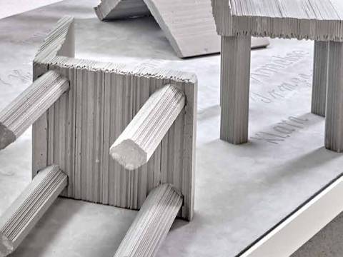 Bilddetail des Denkmals „Never Forget“ – Graue, umgestürzte Stühle und Tisch aus Wellpappe auf hellem Boden. Strukturiertes Material erkennbar. Keine weiteren Elemente sichtbar.