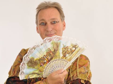  – Person mit blondem Haar haelt einen bunten, dekorativen Faecher mit historischen Szenen und Spitze vor dem Gesicht, laechelt in die Kamera, traegt Kostuem mit