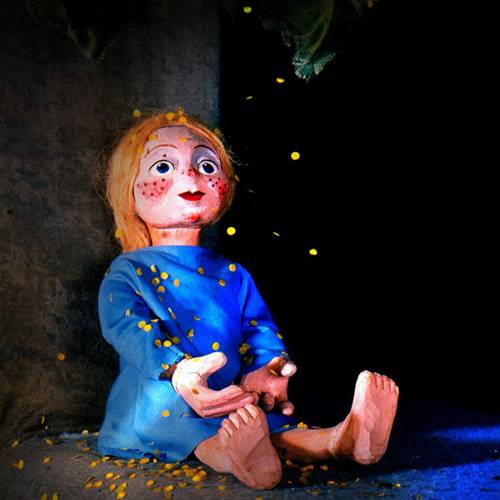 Puppe in blauem Kleid sitzt mit ausgestreckten Beinen, verstreute gelbe Papierschnipsel, schwarzer Hintergrund.