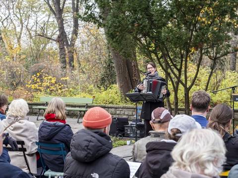  – Person spielt Akkordeon vor Publikum im Freien, umgeben von Bäumen. Schild mit Text: Friedhof der Märtyrer von 1848. Herbstliche Atmosphäre.