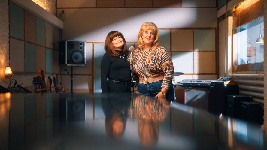 Zwei Personen stehen in einem Aufnahmestudio vor Mikrofonen, umgeben von Musikinstrumenten und Technik. Die Stimmung ist freundlich und professionell.