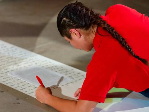  – Eine Person in rotem T-Shirt schreibt mit rotem Stift auf ein Blatt Papier, das auf dem Boden liegt. Auf dem Papier ist handgeschriebener Text.