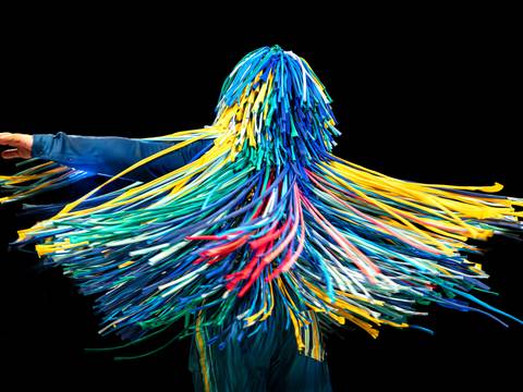 The Sticky Dance – Person mit buntem Fransenanzug in Blau, Gelb, Grün, Rot in Bewegung, schwarzer Hintergrund.