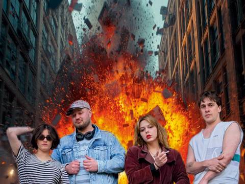  – Vier Personen stehen vor einer Explosion in einer Straßenschlucht; Trümmer und Flammen steigen empor; zwei Wolkenkratzer flankieren die Szene.