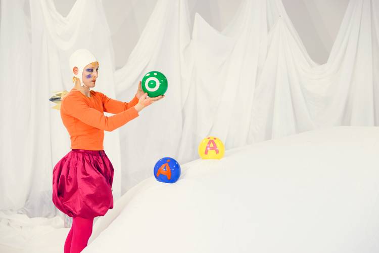 ABC – Das Maler-Alphabet – Person in orangem Oberteil und pinkem Rock haelt gruene Kugel. Gesicht bunt bemalt. Hinter weissem Stoffhuegel liegen zwei Kugeln mit Buchstaben A und O.
