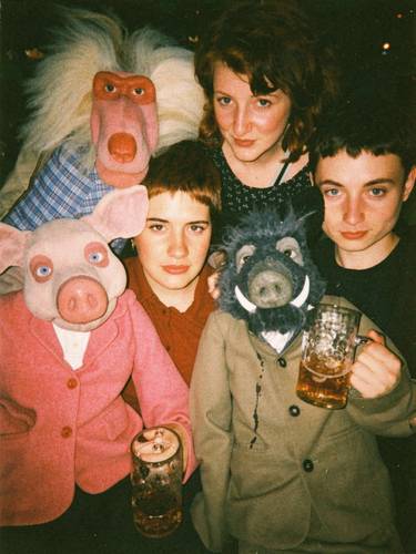 Drei Personen und drei Handpuppen in bunter Kleidung, zwei Bierkrüge. Eine Person hinter den Puppen, eine links, eine rechts, alle schauen in die Kamera.