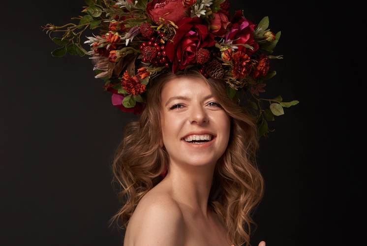 Person mit langem, gewelltem Haar. Großer Blumenkranz aus roten und orangefarbenen Blumen mit grünem Laub auf dem Kopf. Lächelnder Ausdruck.