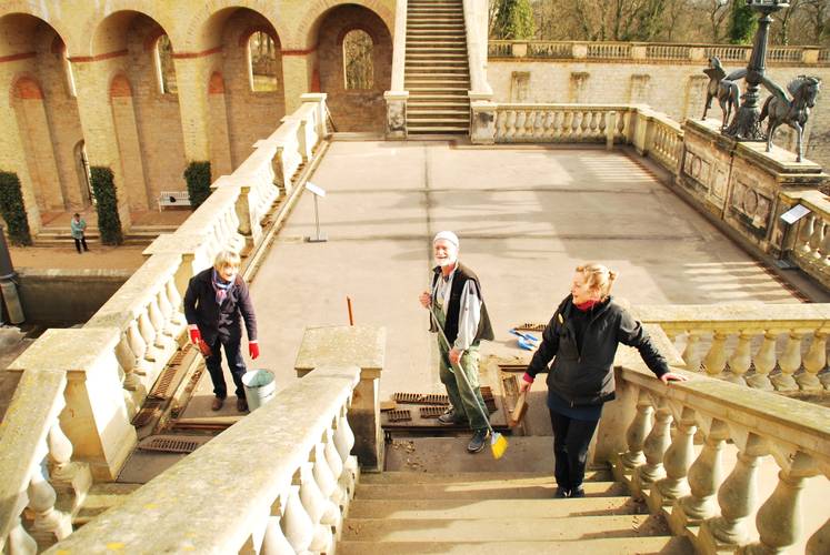 Putzen im Belvedere – Drei Personen reinigen eine Treppe mit Besen und Eimer in einem steinernen Innenhof mit Arkaden und Statuen.