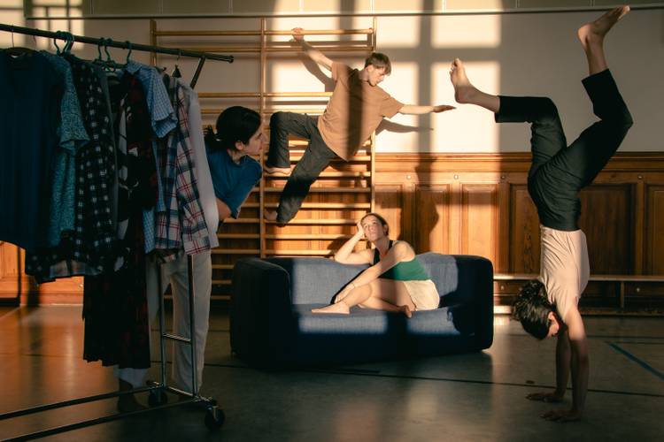 Where Do We Meet – Vier Personen in einem Raum mit Holzsprossenwand und blauem Sofa; eine Person steht auf Händen, eine sitzt, eine hängt an der Wand, eine lehnt an Kleidung.