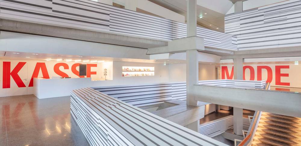 Esther Stocker, ohne Titel, 2023 – Modernes Innenraumdesign mit mehrstöckigen Galerien und großen roten Buchstaben an weißen Wänden: "KASSE" links und "MODE" rechts.