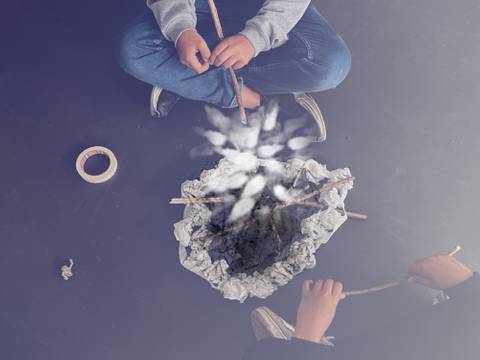  – Zwei Personen sitzen am Boden, halten Stöcke und blicken auf einen weißen Kreis aus zerknülltem Papier. Daneben liegt Klebeband.