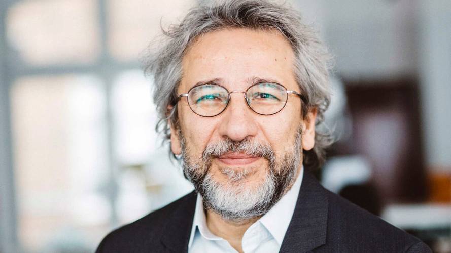 Eine lächelnde Person mit grauen Haaren, Bart und Brille in einem dunklen Blazer, weißem Hemd vor unscharfem Hintergrund.