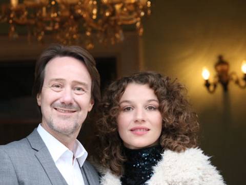 Rainer Bielfeldt und Antonia Bill – Rainer Bielfeldt und Antonia Bill stehen eng nebeneinander und lächeln in die Kamera. Rainer Bielfeldt links trägt ein hellgraues Sakko und ein weißes Hemd, Antonia Bill rechts hat schulterlange lockige Haare, einen dunklen Rollkragen und einen hellen Fellmantel. Im Hintergrund sind ein Kronleuchter und Wandleuchten in einem Innenraum zu sehen.