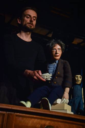 Wohl dem, der keine Heimat hat – Person hält Puppe mit grauen Haaren und braunem Sakko; Puppe sitzt auf Holzmöbel. Im Hintergrund zweite Puppe mit langem dunklem Haar.