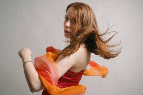 Laura Grandy – Laura Grandy mit langen rötlich-braunen Haaren in einem roten trägerlosen Kleid, umgeben von einem orangefarbenen, fließenden Stoff, der sich durch die Bewegung um sie legt. Sie blickt über die Schulter in die Kamera vor einem hellen, schlichten Hintergrund.