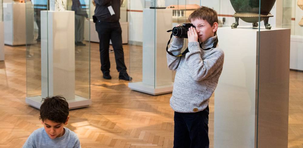 Kind nimmt in einem Museum mit einer Kamera ein Foto auf. Zweites Kind sitzt am Boden. Hinter ihnen Personen und Ausstellungsstücke in Glasvitrinen.