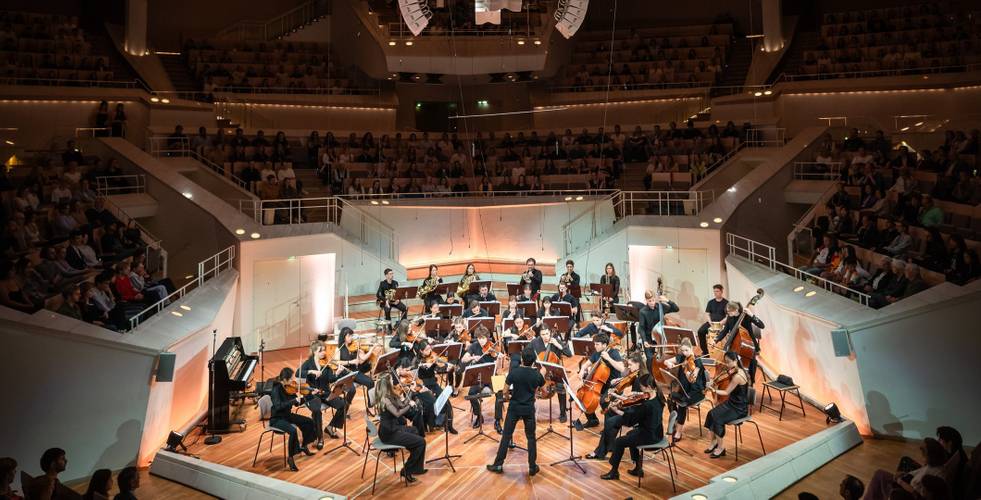 Person dirigiert Orchester auf Bühne, umgeben von Publikum in modernem, großen Konzertsaal. Musiker*innen spielen verschiedene Streichinstrumente.
