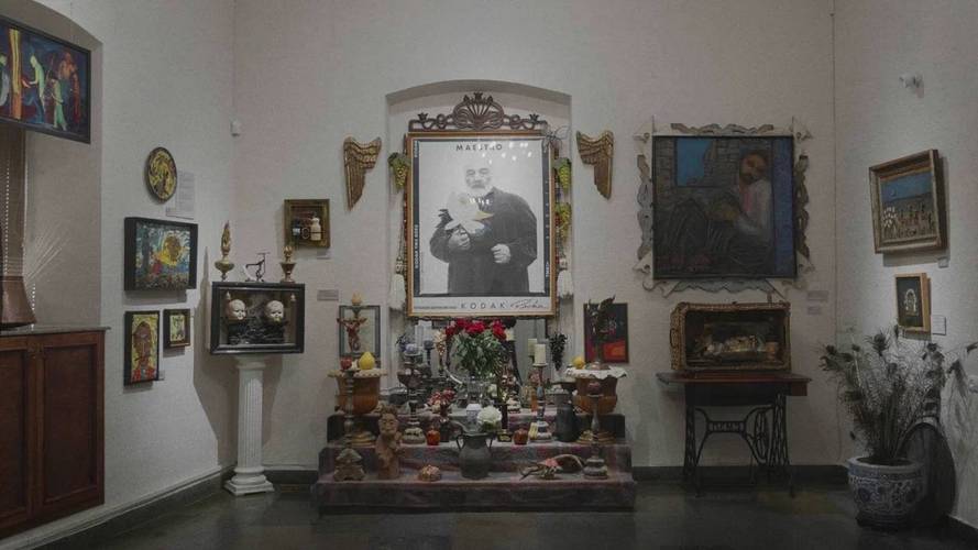 Kunstgalerie mit mehreren Gemälden, einer zentralen Fotografie einer Person mit Hund, dekorativen Objekten und Blumen auf einem Tisch darunter.