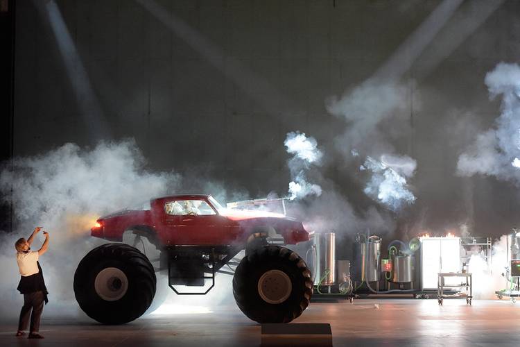 Die Entführung aus dem Serail – Roter Monstertruck mit Rauch und Lichteffekt; Person fotografiert. Im Hintergrund industrielle Metallstrukturen mit Lichtquellen.