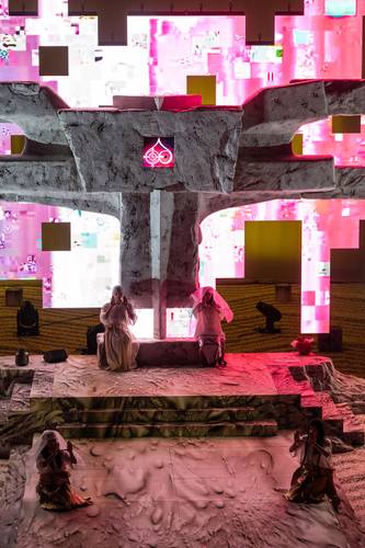 Mittwoch aus Licht – Drei Personen in weißen Gewändern vor einer großen Steinskulptur mit pinkem Licht. Hintergrund mit lebhaften rosa und gelben Quadraten.