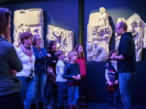 Familienführung in Pergamon. Das Panorama – Personengruppe betrachtet antike Reliefs in einem Museum. Eine erwachsene Person spricht zu den anderen, darunter mehrere Kinder und Erwachsene.