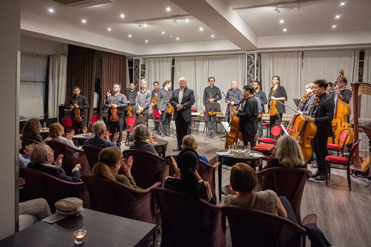 Eine Gruppe von Musiker*innen mit Streichinstrumenten steht verneigend vor sitzendem Publikum in einem Raum mit roten Stühlen und hellen Vorhängen.