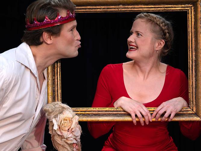 Märchen im Glaspalast von und mit dem Hexenberg Ensemble: Rapunzel – Männlich gelesene Person mit roter Krone küssend, weiblich gelesene Person im roten Kleid lächelnd, beide halten einen goldenen Rahmen.
