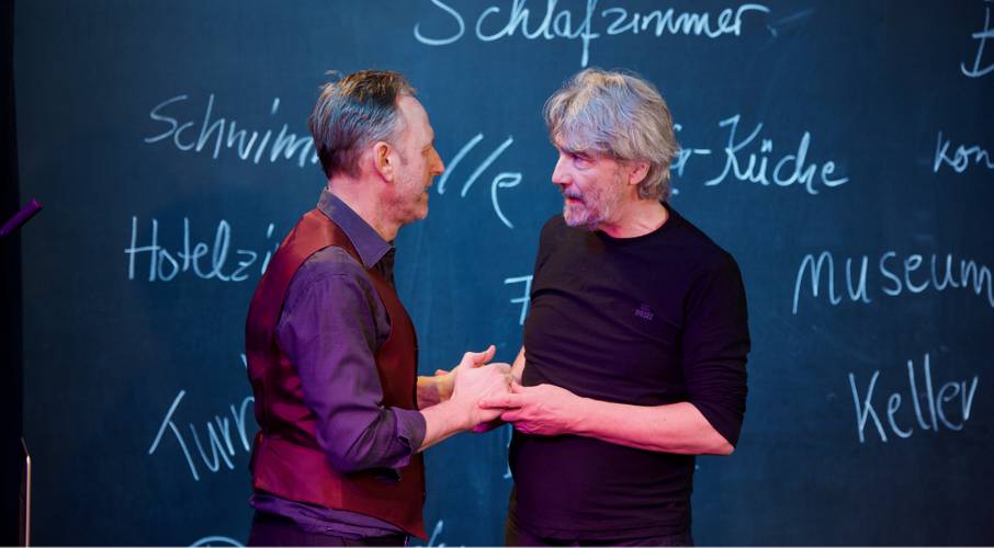 Zwei Personen stehen vor einer Tafel mit den Woertern Schlafzimmer, Schwimmbad, Hotelzimmer, Museum, Keller. Die Personen halten miteinander Haende.