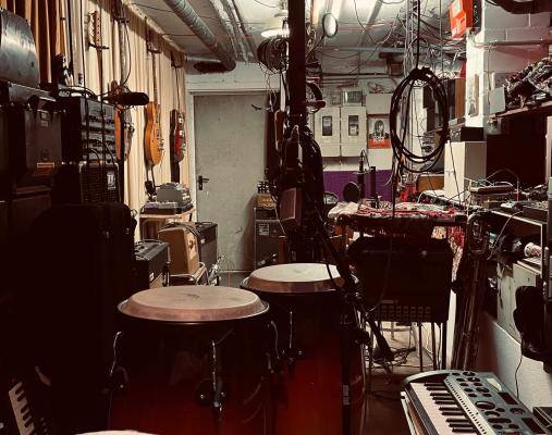 Stroum – Musikraum mit Congas, Keyboards, Kabeln, Verstärkern, Gitarren an der Wand, kleinem Tisch, Regal und Mischpulten. Dezente Beleuchtung.