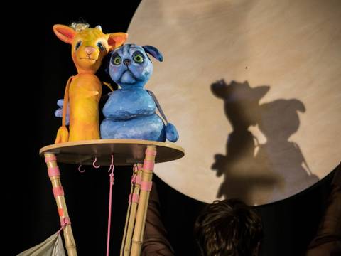 Das große Wohin – Zwei bunte Marionetten, ein orange Tier und ein blaues Tier, stehen auf einer Plattform vor einer grossen, runden, beleuchteten Scheibe.