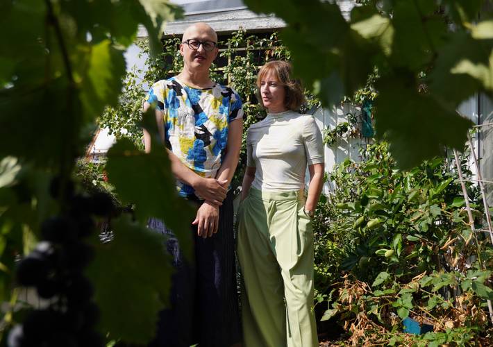 Zwei Personen stehen in einem Garten, umgeben von Pflanzen. Eine trägt ein bunt gemustertes T-Shirt und eine Brille, die andere eine hellgrüne Hose und ein
