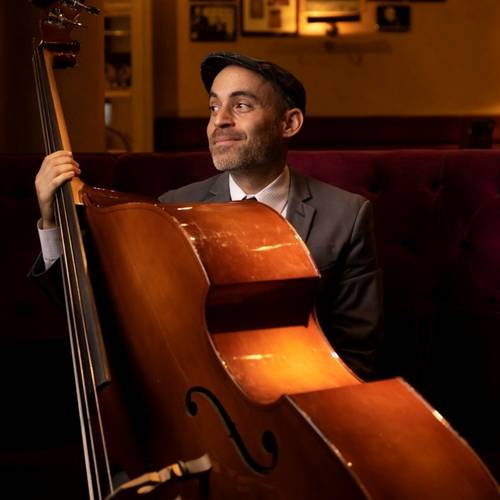 Tal Ronen – Person mit Cello in edlem Ambiente, goldene Beleuchtung; trägt Anzug, Hemd und Schiebermütze, schaut lächelnd zur Seite, lila Samtsofa im Hintergrund.