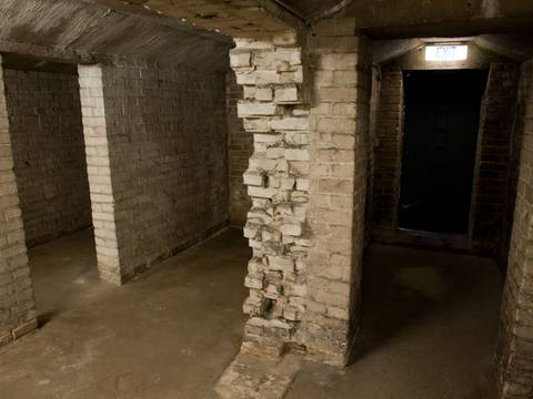 Keller der "Baracke 13" – Unterirdischer Raum mit grob verputzten Ziegelwänden, mehreren Pfeilern und einem beleuchteten Ausgangsschild über einer offenen Tür.