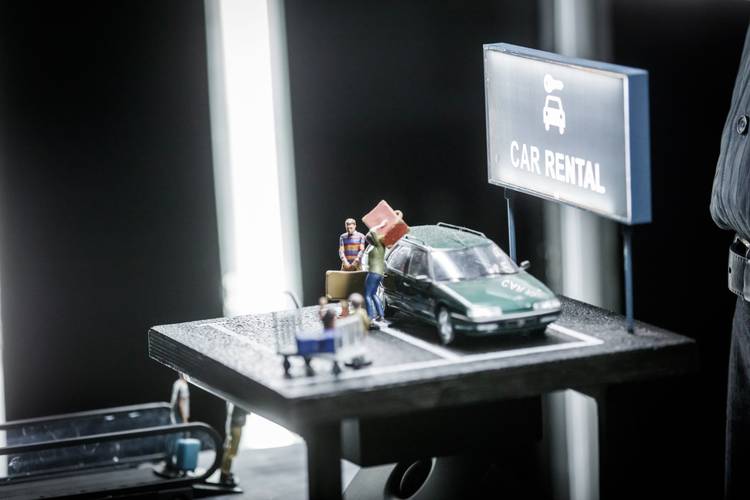 Transport: Connecting Flights – Miniaturautoszene mit grünem Auto, Figur mit Koffer, Schild "CAR RENTAL", beleuchteter Hintergrund, dunkler Rahmen, Modellcharakter.