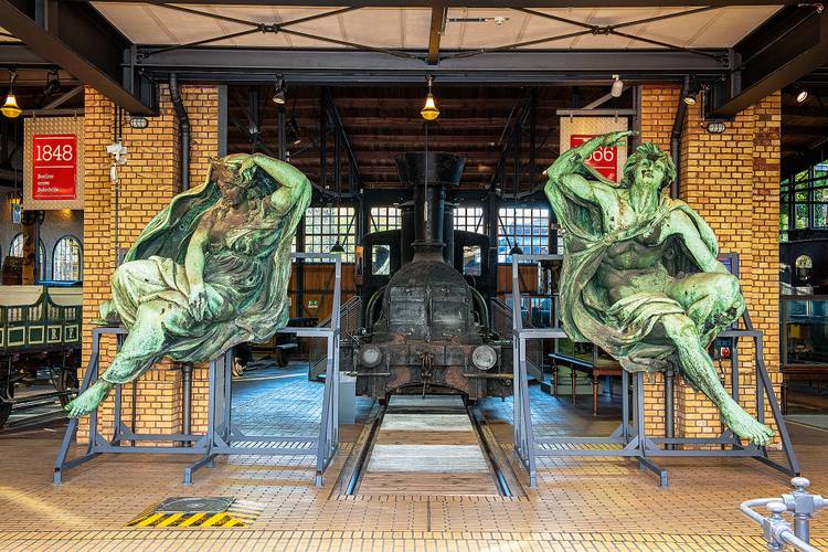 Die Skulpturen „Tag“ und „Nacht“ des Bildhauers Ludwig Brunow (1843-1913) befanden sich einst über dem Haupteingang des Anhalter Bahnhofs. Heute begrüßen die restaurierten Galvanoplastiken die Besucherinnen und Besucher am Eingang zu den Lokschuppen. – Zwei große, grün angelaufene Bronze Figuren stehen links und rechts vor den gelben Backsteinsäulen der historischen Lokschuppen. Zwischen Ihnen steht eine schwarze Lokomotive.