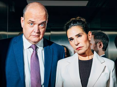  – Zwei Personen in Anzug und Blazer stehen nebeneinander in einem Aufzug, der metallene Wände hat.