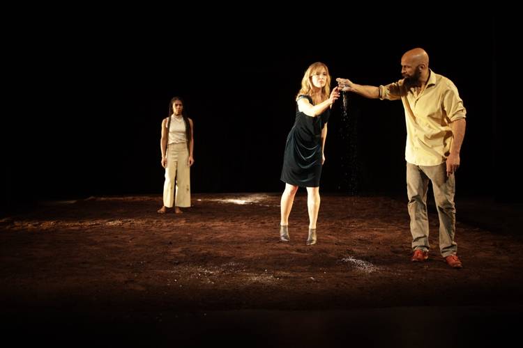 Aesthetic of Color – Zwei Personen stehen auf dunkler Erde. Eine Person gießt Erde in die Hand der anderen. Im Hintergrund steht eine dritte Person und schaut zu.
