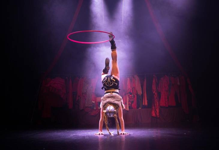 Barbaren Barbies: A wild women circus – Person balanciert auf Haenden, ein pinker Reifen schwebt ueber dem Kopf. Hintergrund mit bunten Kostuemen beleuchtet. Bühne in Nebel gehüllt.