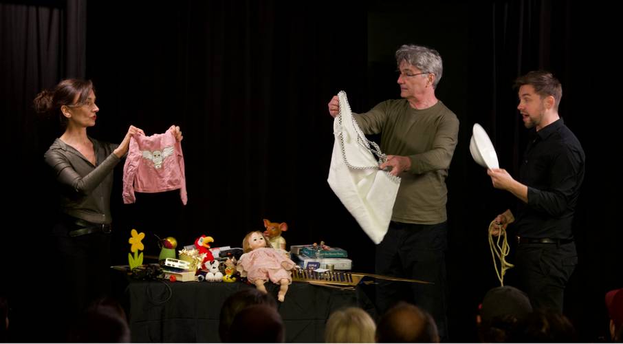 Drei Personen auf einer Bühne, eine mit rosa Kinderpullover, eine mit weißem Stoff und eine mit weißem Teller. Tisch mit Spielzeug und Puppe. Theaterkulisse.