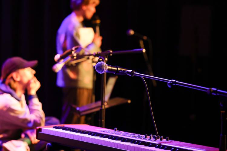 Im Vordergrund ist ein Mikrofon zu sehen, das über einem E-Piano positioniert ist. Im dunklen Hintergrund sind zwei Personen zu erkennen, von denen eine sitzt und die andere ein Mikrofon in der Hand hält.