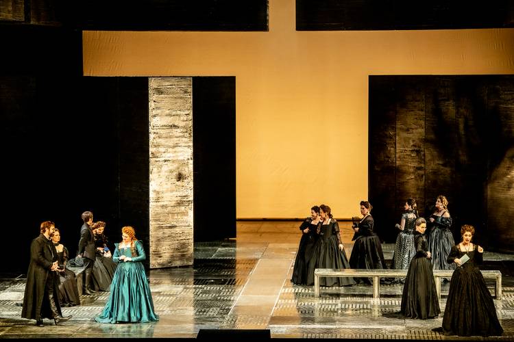 Don Carlo – Mehrere Personen in historischen Kostümen auf einer Bühne mit großen dunklen Wänden und einem gelben Hintergrund. Zwei Gruppen stehen links und rechts.