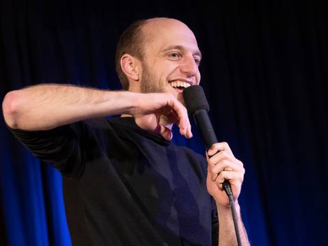 Porträt des Autors und Poetry-Slam-Meisters Alex Burkhard – Eine lachende Person hält ein Mikrofon in der rechten Hand, trägt ein schwarzes Langarmshirt und steht vor einem dunklen Vorhang.