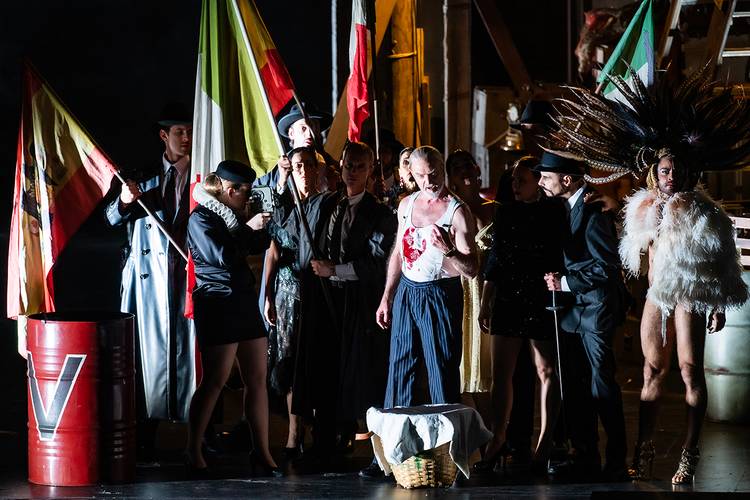 La forza del destino – Eine Gruppe von Personen in bunter Kleidung, eine trägt Federschmuck, hält Nationalflaggen, steht bei Metallfass mit rotweißem "V" im Theater.