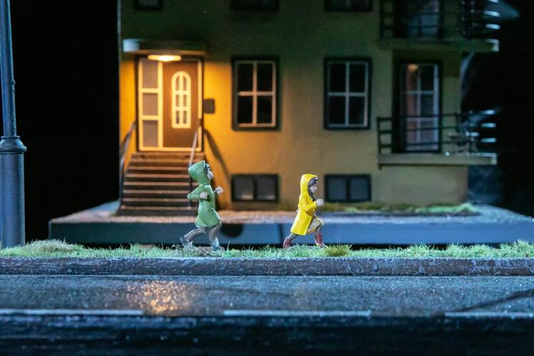 Transport: Departure – Zwei Figurinen in Regenmänteln auf einem nassen Bürgersteig vor beleuchtetem Haus; die linke Figur trägt grün, die rechte gelb mit roten Stiefeln.