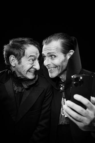 Tonio Arango und Boris Aljinović sind Dracula und sein Lakai Igor. – Zwei Personen lächeln einander an, in dunkler Kleidung mit Krägen. Eine Person hält ein Smartphone und macht ein Selfie. Schwarz-weißes Bild.