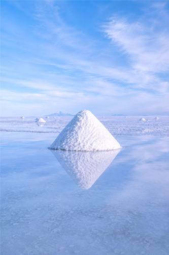Ein Salzberg pyramidenförmig in spiegelnder Salzebene. Wolkiger Himmel oben. Mehrere kleinere Salzberge im Hintergrund.