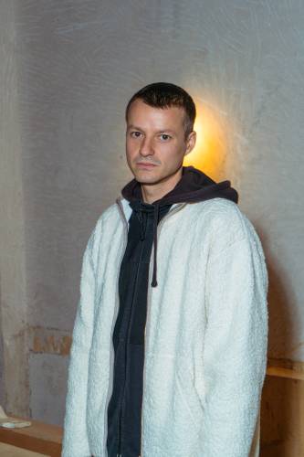 Person trägt weißen Flauschpullover und schwarze Jacke. Hintergrund aus beige strukturiertem Beton mit gelbem Licht.