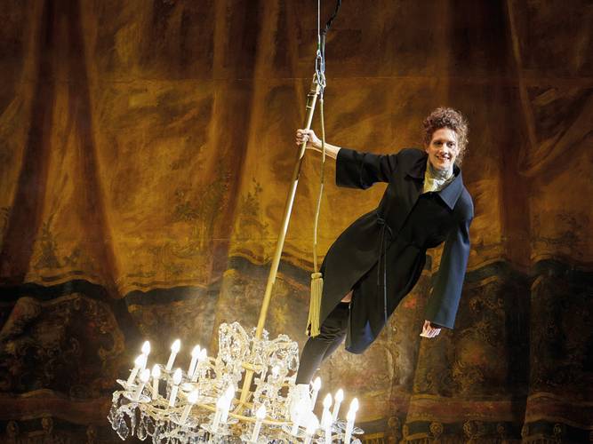 Fräulein Else – Eine Frau in einem Blazermantel steht lächelnd auf einem schwingenden Kronleuchter