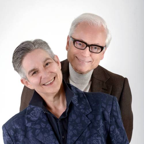 Sigrid Grajek und Hans-Jürgen Schatz – Zwei lächelnde Personen; die vordere trägt ein florales Sakko, die hintere eine Brille und einen Pullover unter einem Sakko.