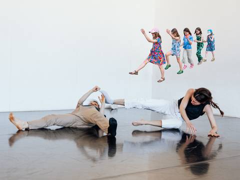  – Zwei Personen liegen am Boden eines Tanzstudios. Fünf Kinder in bunten Kleidern schweben in der Luft, als ob sie hüpfen.